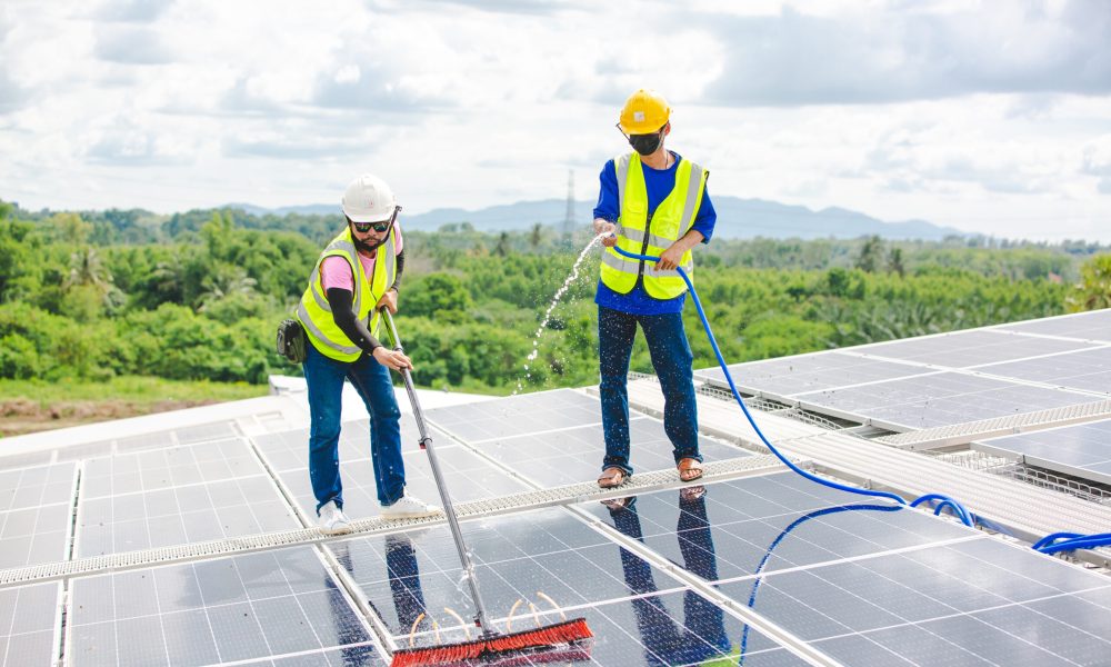 Professional worker cleaning solar panels with brush and washing with water on roof structure of building factory. Technician using mop to clean the dirty and dust, green electricity energy technology